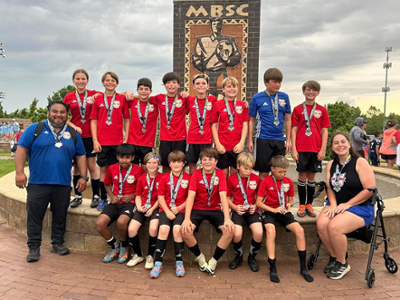 Youth soccer team celebrates victory. Players and coaches, including one seated on a walker, pose together wearing medals. All smiling proudly.